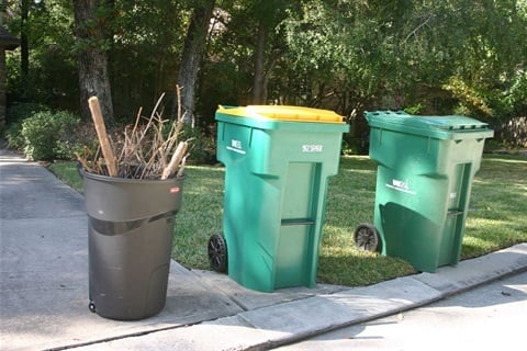 Three waste containers on a sidewalk: a gray bin with branches and sticks on the left, and two green recycling bins on the right, with trees in the background